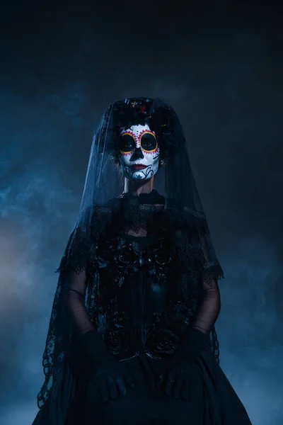 Front view of woman in scary halloween makeup and black costume with veil on dark blue foggy background — Stock Photo