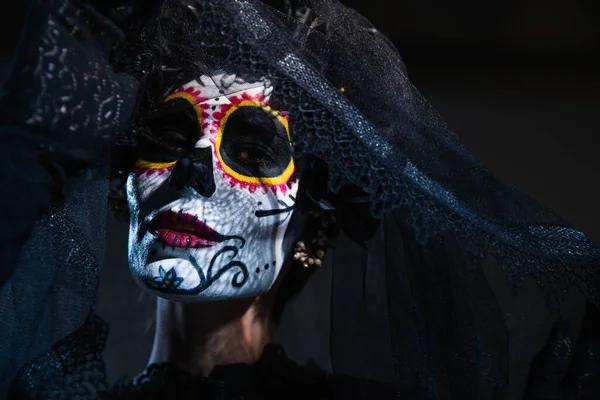 Portrait de femme avec crâne de sucre halloween maquillage regardant caméra près voile de dentelle noire sur fond bleu foncé — Photo de stock