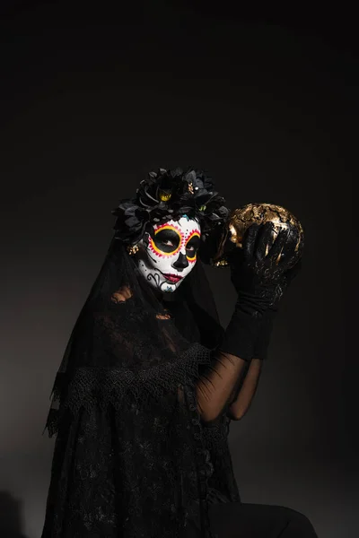 Woman with traditional santa muerte makeup and costume with dark wreath holding golden skull on black background — Stock Photo
