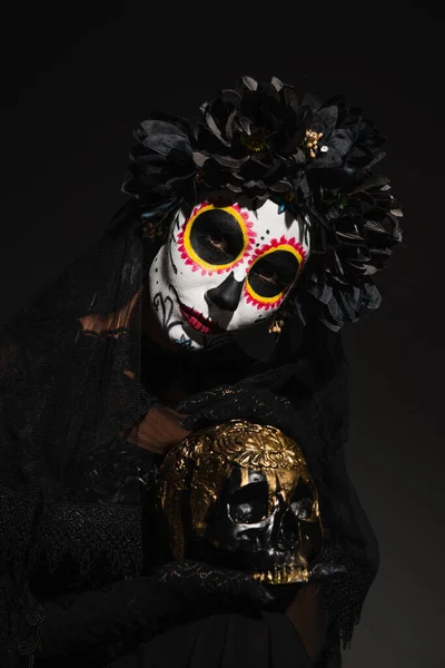 Frau in dunklem Kranz und gespenstischem Halloween-Make-up mit goldenem Totenkopf auf schwarzem Hintergrund — Stockfoto