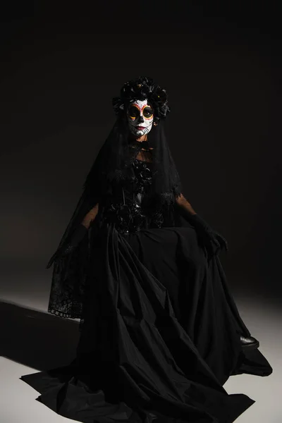 Longitud completa de la mujer en vestido negro con velo y maquillaje de Halloween espeluznante sentado sobre fondo negro - foto de stock