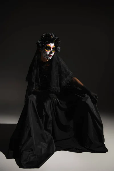 Longitud completa de la mujer en vestido de bruja negro y espeluznante maquillaje de halloween sentado sobre fondo oscuro - foto de stock