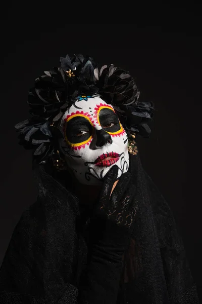 Woman in dark wreath touching face with traditional santa muerte makeup looking at camera isolated on black — Stock Photo