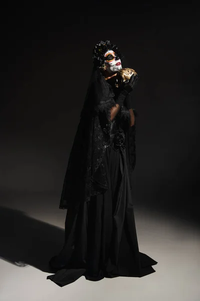 Full length of woman in black witch costume and traditional santa muerte makeup standing with golden skull on dark background — Stock Photo