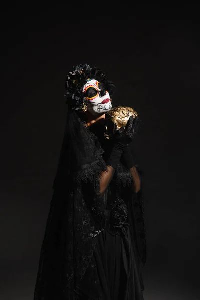 Frau mit Zuckerschädel-Make-up trägt dunkles Kostüm mit Schleier und hält goldenen Totenkopf isoliert auf schwarz — Stockfoto