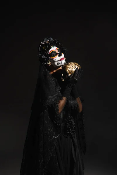 Mujer con maquillaje de cráneo de azúcar usando traje oscuro con velo y sosteniendo cráneo dorado aislado en negro - foto de stock