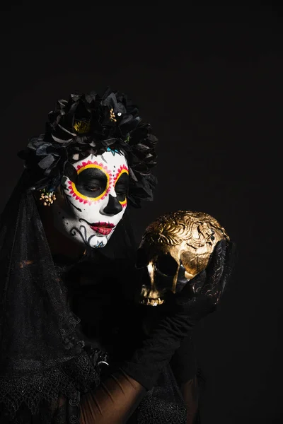 Mujer en traje de halloween oscuro y maquillaje espeluznante celebración de cráneo de oro aislado en negro - foto de stock