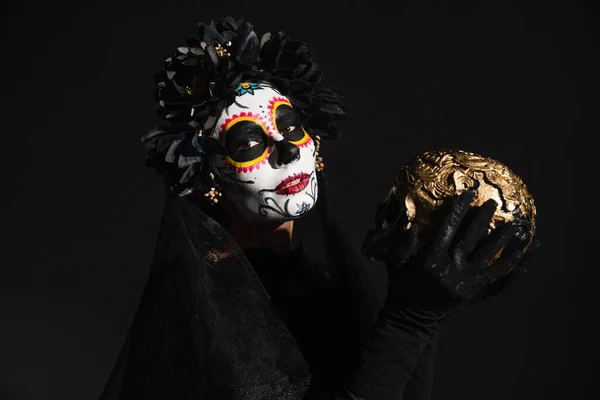 Mujer en maquillaje de cráneo de azúcar y traje oscuro espeluznante celebración de cráneo de oro aislado en negro - foto de stock