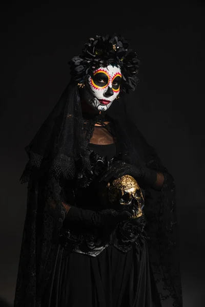 Mujer en traje espeluznante y maquillaje catrina celebración de cráneo de oro aislado en negro - foto de stock