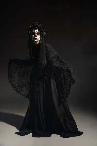 Piena lunghezza della donna in costume da Santa Muerte spettrale e trucco catrina su sfondo scuro — Foto stock