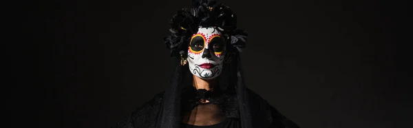 Portrait of woman in sugar skull makeup and dark wreath looking at camera isolated on black, banner — Stock Photo