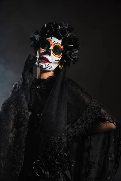 Frau in Halloween-Make-up und Hexenkostüm mit schwarzem Spitzenschleier, stehend mit der Hand auf der Taille auf dunklem Hintergrund — Stockfoto