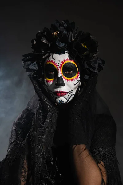 Woman in black wreath holding hands near face with sugar skull makeup on dark smoky background — Stock Photo