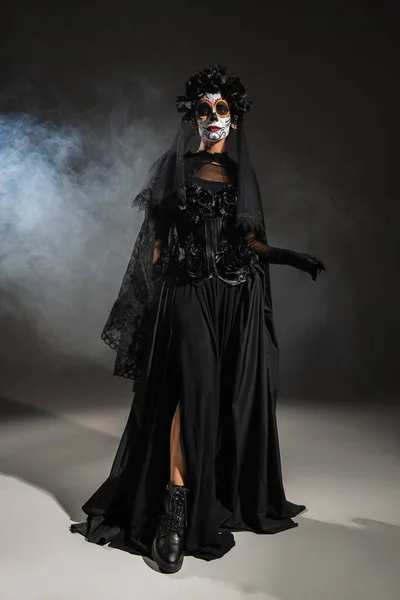 Full length of woman in black halloween costume with veil and spooky makeup on dark background with smoke — Stock Photo
