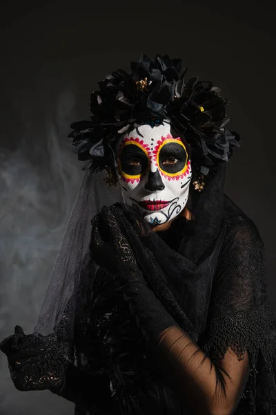 Woman in black wreath and traditional santa muerte makeup looking at camera on dark foggy background — Stock Photo