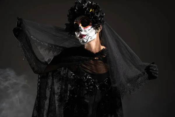 Woman in spooky halloween makeup and witch costume holding black lace veil on dark background with smoke — Stock Photo