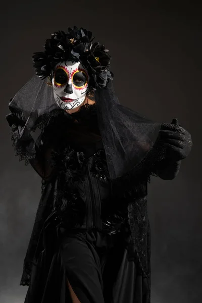 Frau in Halloween-Zuckerschädel-Make-up und schwarzem Kostüm mit Schleier blickt auf dunklem Hintergrund in die Kamera — Stockfoto