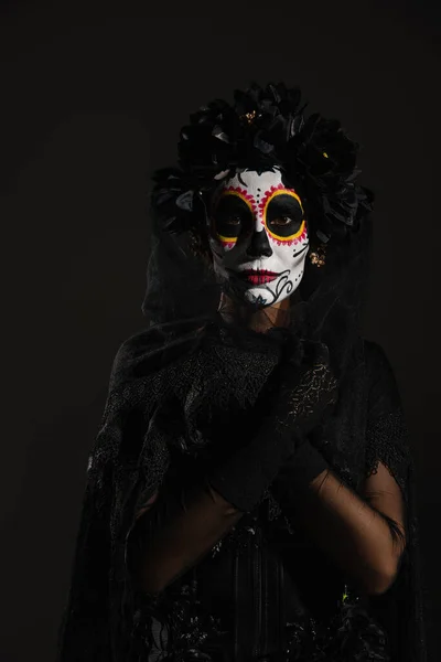 Woman with mexican day of dead makeup wrapping herself in dark veil and looking at camera isolated on black — Stock Photo