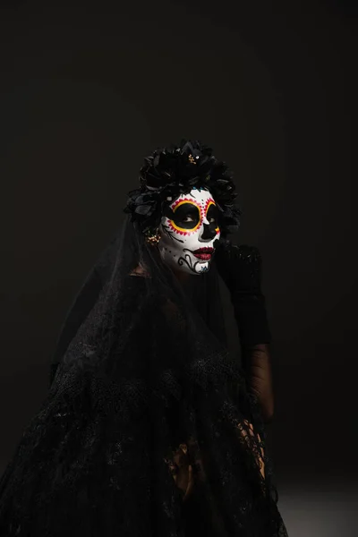 Mujer en espeluznante maquillaje de halloween y corona negra con velo mirando a la cámara sobre fondo oscuro - foto de stock