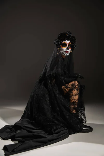 Повна довжина жінки з макіяжем з цукрового черепа в чорній сукні з мереживною вуаллю, що позує на темному фоні — стокове фото