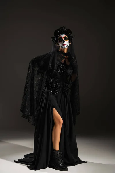 Full length of woman in sugar skull makeup posing in black halloween costume on dark background — Stock Photo