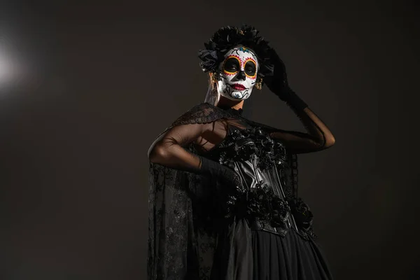 Low angle view of woman in sugar skull makeup and halloween costume posing with hand on hip on black background — Stock Photo