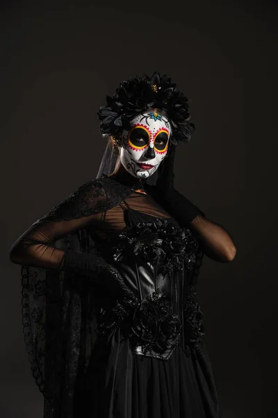 Femme en costume d'Halloween sombre et maquillage du crâne de sucre posant à la main sur la taille isolé sur noir — Photo de stock