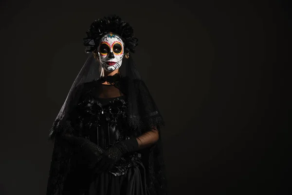 Mujer en traje oscuro y aterrador y maquillaje de cráneo de azúcar de pie aislado en negro - foto de stock