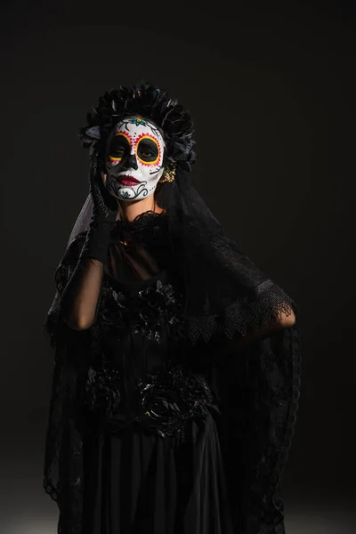 Жінка в мексиканському макіяжі Катріна і темний костюм з мереживною завісою позує з рукою на стегні на чорному тлі — стокове фото
