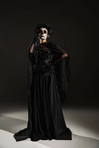 Longitud completa de la mujer en vestido de bruja negro y el maquillaje de Halloween miedo posando con la mano en la cadera sobre fondo oscuro - foto de stock