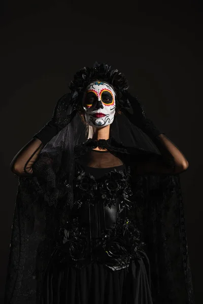 Mulher em maquiagem catrina e traje de santa muerte tocando grinalda escura com véu isolado em preto — Fotografia de Stock