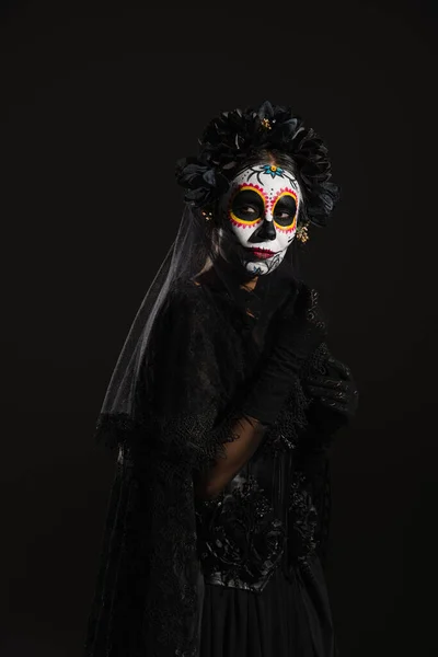 Woman in sugar skull makeup and dark halloween costume with veil looking away isolated on black — Stock Photo