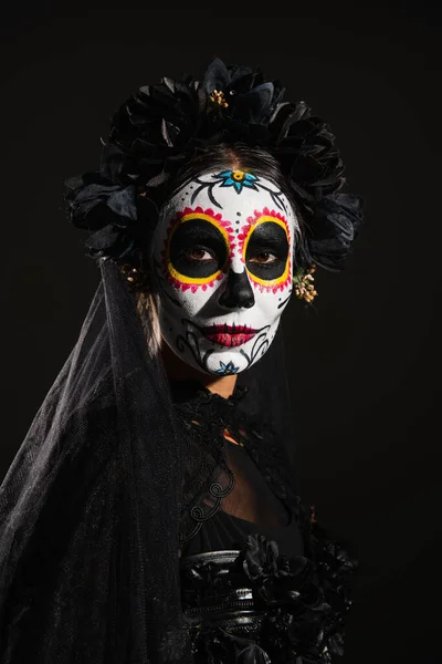 Retrato de mujer con espeluznante maquillaje de halloween y velo oscuro mirando a la cámara aislada en negro - foto de stock