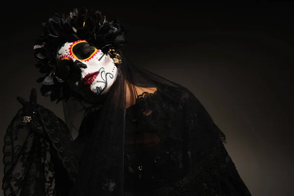 Mujer en espeluznante maquillaje de halloween y corona oscura con velo de encaje posando con los ojos cerrados sobre fondo oscuro - foto de stock