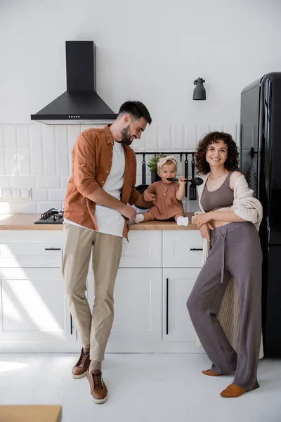 Младенец в повязке держит ковш у рта, сидя на кухонном столе рядом с счастливыми родителями — стоковое фото