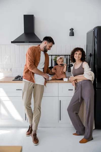 Bébé fille dans bandeau tenant louche près de la bouche tout en étant assis sur le plan de travail de la cuisine près de parents heureux — Photo de stock