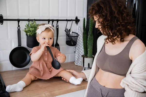 Bébé fille dans bandeau tenant louche près de la bouche tout en étant assis sur le plan de travail de la cuisine près de mère heureuse — Photo de stock