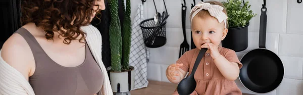 Niña en diadema sosteniendo cucharón cerca de la boca mientras está sentado en la encimera de la cocina cerca de la madre rizada, pancarta - foto de stock