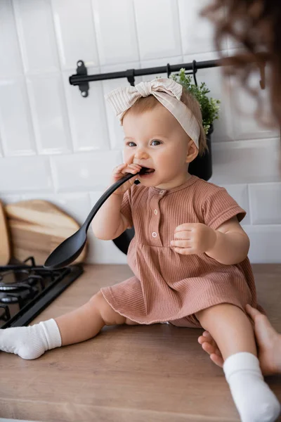 Младенец в повязке держит ковш у рта, сидя на кухонном столе рядом с матерью — стоковое фото