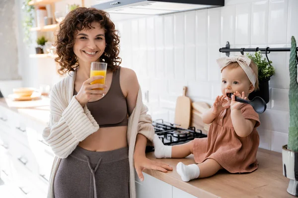 Bébé fille tenant louche près de la bouche tout en étant assis sur le plan de travail de la cuisine près de mère heureuse avec un verre de jus d'orange — Photo de stock