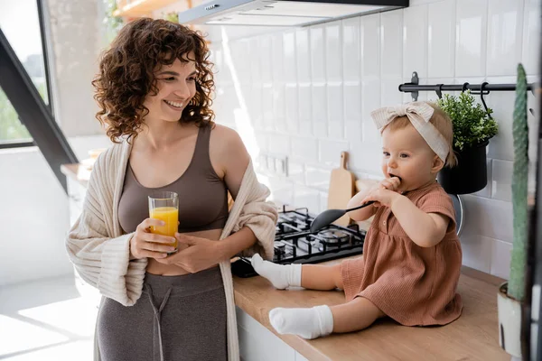 Новорожденная девочка держит ковш у рта, сидя на кухонном столе рядом с веселой матерью со стаканом апельсинового сока — стоковое фото