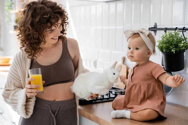 Девочка сидит на кухонном столе рядом с веселой матерью со стаканом апельсинового сока и мягкой игрушкой — стоковое фото