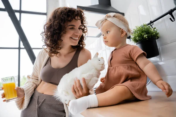 Infant girl sitting on kitchen worktop near positive mother with glass of orange juice and soft toy — Stock Photo