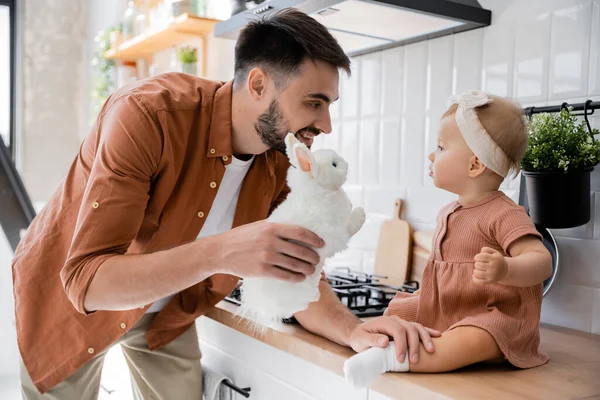 Счастливый бородатый мужчина держит мягкую игрушку рядом с младенческой дочерью, сидя на кухонном столе — стоковое фото