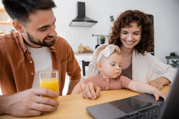 Alegre hombre sosteniendo vaso de jugo de naranja cerca de esposa e hija lactante utilizando el ordenador portátil - foto de stock