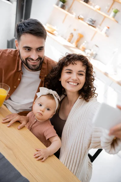Curly woman taking selfie with cheerful husband and infant daughter at home — Stock Photo