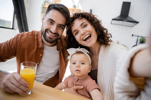 Donna riccia sorridente vicino marito allegro e figlia neonata mentre guarda la fotocamera — Foto stock