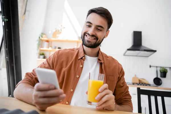 Cheerful and bearded man holding glass of orange juice while using smartphone — Stock Photo