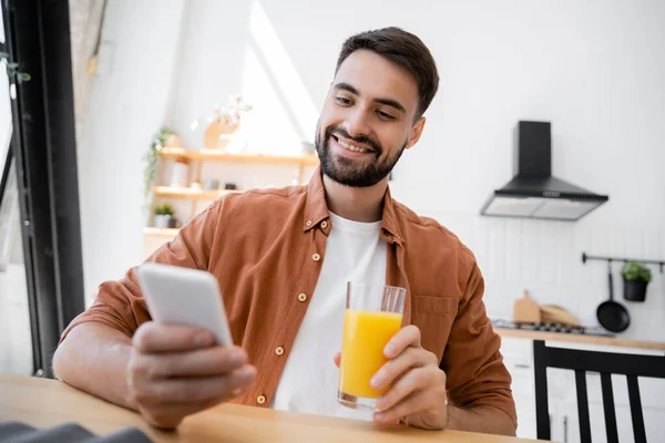 Homem alegre e barbudo segurando vidro de suco de laranja ao usar o smartphone — Fotografia de Stock