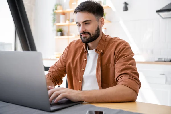 Freelancer barbudo usando laptop enquanto trabalhava em casa perto de smartphone na mesa — Fotografia de Stock